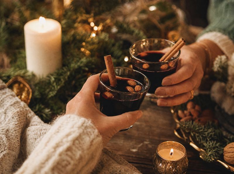 Händer som håller glögg med kanelstänger intill levande ljus och granris – inspirationsbild